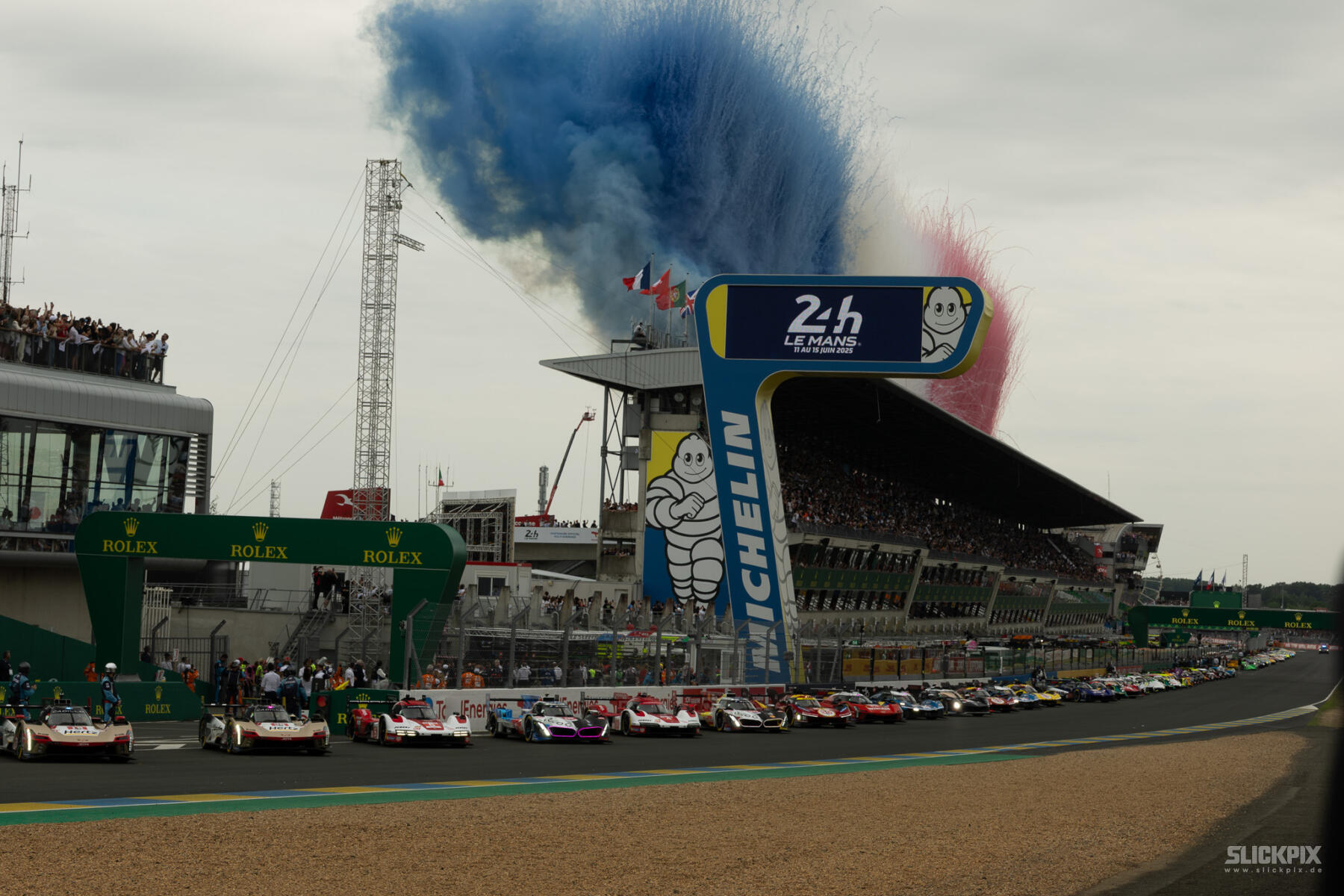Die Rennwagen stehen am Start der 24 Stunden von Le Mans, während blauer und rosa Rauch in den Himmel aufsteigt und im Hintergrund die Tribünen voller Zuschauer zu sehen sind.